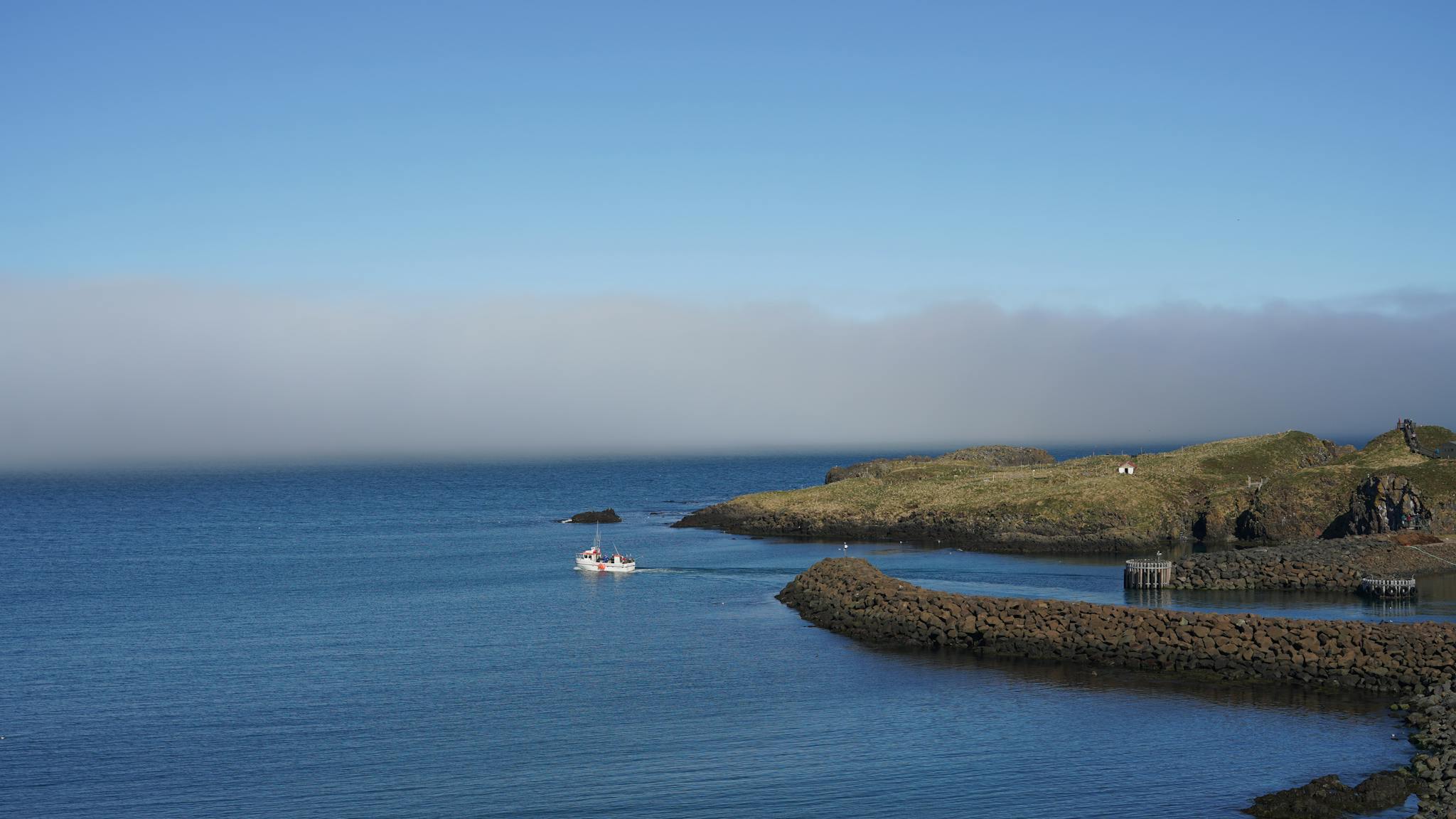 A tranquil scene of a boat navigating calm waters near rocky Icelandic coast under a clear blue sky.