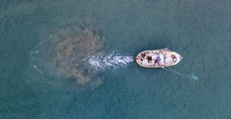 From above of unrecognizable person floating on small motorboat with sailing equipment in rippling blue sea and trying to catch fish