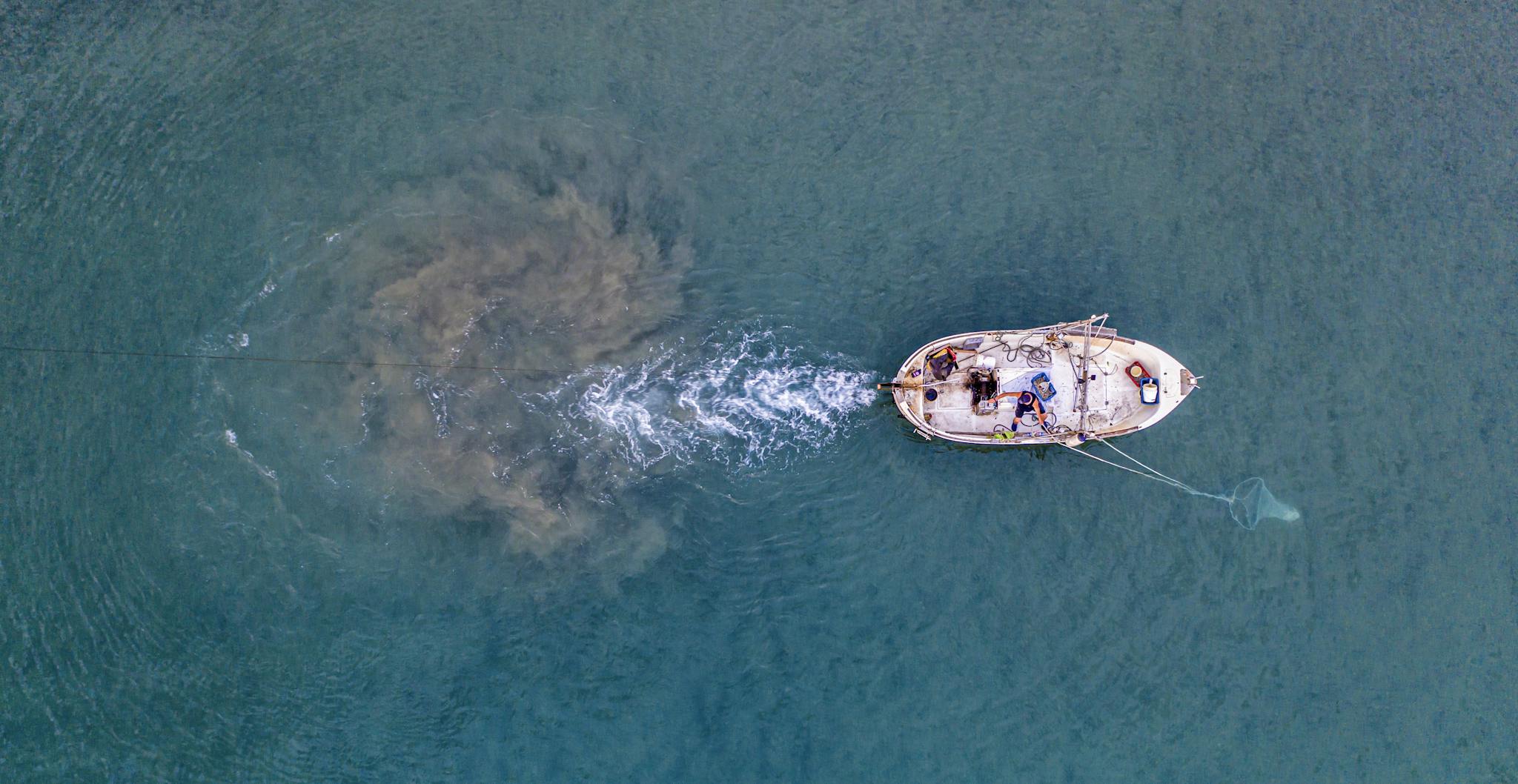 From above of unrecognizable person floating on small motorboat with sailing equipment in rippling blue sea and trying to catch fish